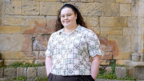 An individual stands in front of a textured stone wall, wearing glasses and a patterned short-sleeve shirt with heart motifs. Curly hair is held back with a dark headband, and the person’s hands are placed casually in pockets. The background features weathered bricks and small patches of greenery near the base.