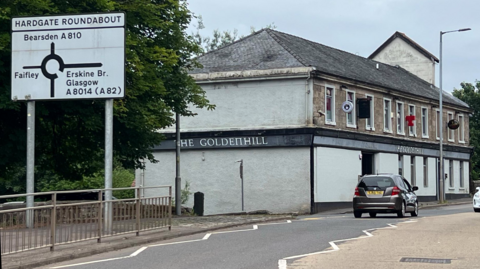 Road with a pub on it. There are cars driving past and a road sign showing that a roundabout is coming up.