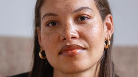 Seren Jones, who is of mixed race and has long brown hair, smiles at the camera. She is wearing gold hoop earrings and has freckles.