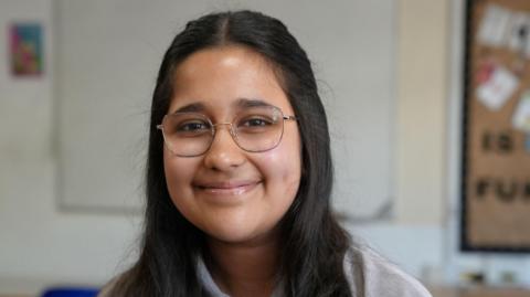 A girl smiling sweetly at the camera. She is wearing thin-framed glasses and has long black hair. She is in a classroom.