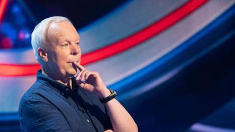 Actor Steve Pemberton wearing a navy short-sleeved shirt appearing on BBC One's The Weakest Link. He has his finger over his mouth as he answers a question. 