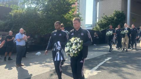 Newcastle United head coach Eddie Howe walks with club players and staff carrying a floral wreath of white and cream flowers with green foliage for Kevin Keegan at St James' Park. Several bystanders clap as he walks by.