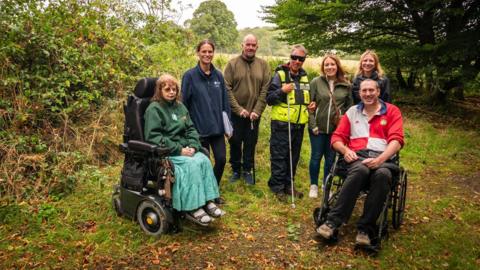 Six people are at a rural farm. Behind them are trees and shrubs.