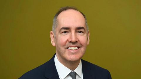 A man with thinning grey hair and wearing a suit, smiling for the camera. The image is cropped off around his shoulders. 