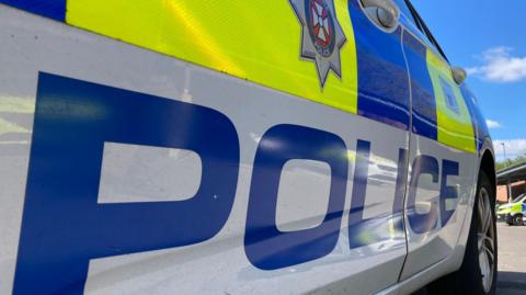 The side of a police vehicle parked in a car park. The word 'police' can be seen on the bottom and the force's logo is on the door.