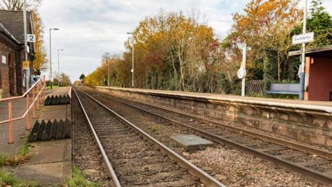 The railway station at Swinderby. A shelter can be seen on one side of the tracks, along with a location sign, and the front of larger building on the other