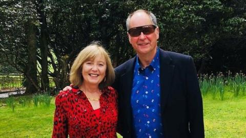 Mary and David Crowley smiling at the camera, she is wearing a red and black dress, he is wearing a dark jacket over a blue shirt and blue jeans.