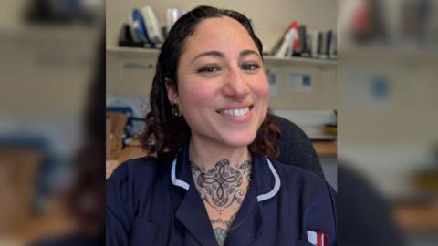 A selfie of Nicole Durman wearing dark navy hospital scrubs and smiling at the camera. She has dark curly hair in a middle parting, a septum piercing, a stretcher ring in her right ear lobe and a large neck tattoo depicting a swirling, symmetrical Celtic pattern. Behind her there is a desk and a shelf with medical files stacked on them.