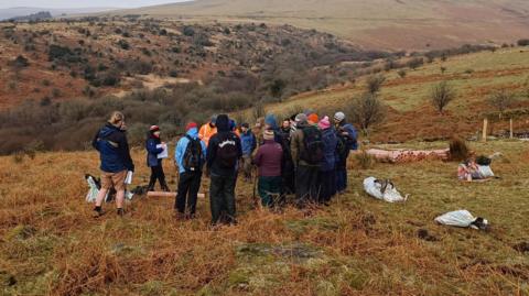 A group of volunteers wearing various wet weather gear stand on Harford Moor in a circle. A person in the centre of the group is pointing at a piece of paper. The moor is green and brown and looks damp. 