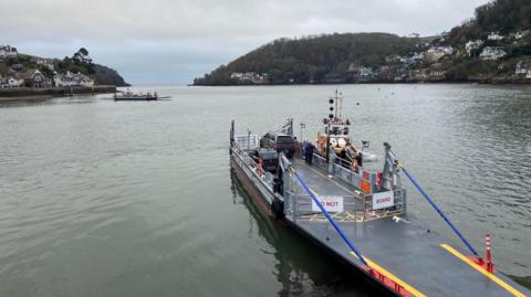 A ferry is seen on the River Dart in Devon. There are two cars on it and it is docked at shore. 