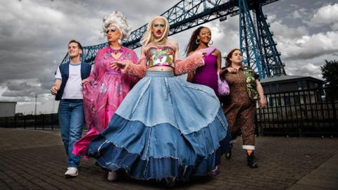 The Smoggie Queens cast lined up walking towards the camera with an industrial structure behind them.
