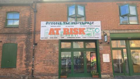 Exterior of a red-brick building with green-framed windows and doors. A large banner reading “Protect The Warehouse - At Risk” hangs above the entrance, promoting a community garden, bike shop, book shop, meeting space, co-working, radio and podcast studio, workshops, and environmental initiatives. 