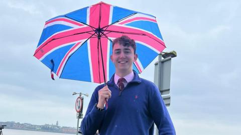 Harry smiles at the camera, he is wearing a blue sweater with a pink shirt and burgundy tie. He is holding an umbrella with a union jack print on it. 