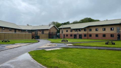 A modern-looking brown building, with a tarmac road leading up to it. In the foreground are areas of grass, with benches 