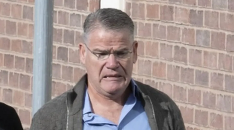 A man with grey hair, glasses and a grey cardigan standing near a brick wall