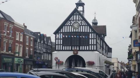 A black and white pitched market hall building in the centre of a high street. Pedestrians can walk through via an archway. Three-storey buildings line the street on either side with shop signs visible. A row of parked car roofs is visible at the foot of the market hall.