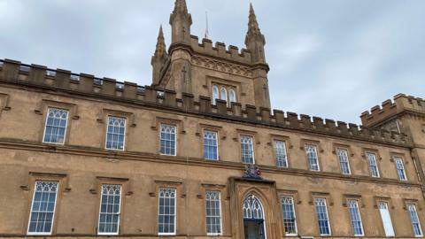 A picture of the front of Elizabeth College. It is a large brown building with multiple windows and a large entrance. It is a castle like building.