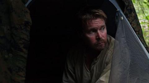 A man with a short light brown beard sits in a tent pitched in a forest clearing. He looks lost in thought.