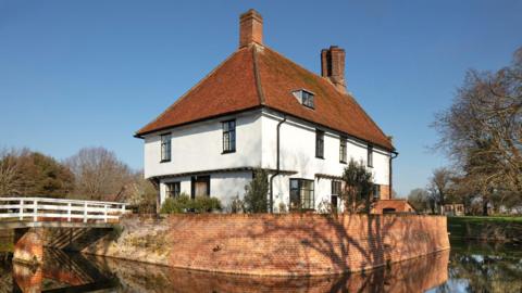 A general view of Letheringham Lodge - a white building with an orange tiled roof surrounded by a moat and trees. 