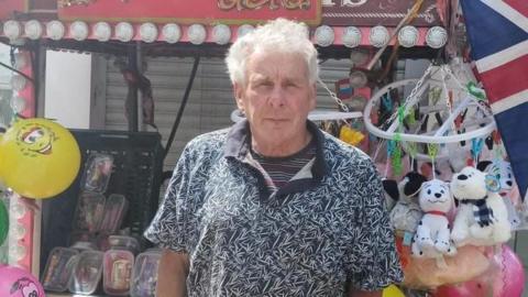 A man with medium-length white hair stands in front of a stall with toys on display.