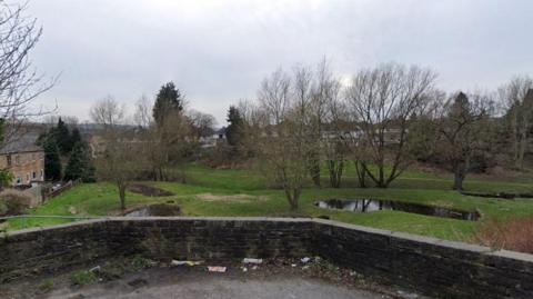 The picture overlooks a green space with several trees and hedges. Two small bodies of water can be seen. To the left, a row of houses stretches.