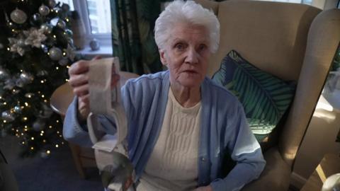 Jean Waddy sits in a beige armchair. She has short grey hair and is wearing a white top and a light blue buttoned sweater. She is looking at the camera and holding up a paperchain decoration made out of newspaper. A Christmas tree covered with silver decorations is lit up on the left.