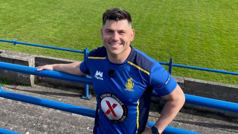 James Newton, smiling and holding on to a blue standing rail at Whitehaven's ground in a blue and yellow Whitehaven training top