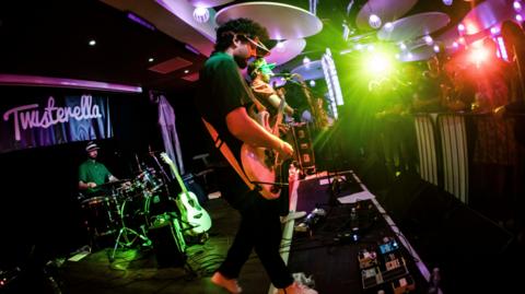 Melin Melyn performing at the Twisterella in 2023. One band member is playing the guitar in the front, while another one next to him is singing.  A third is playing the drums at the back in front of the festival's logo. There are green and red lights. Audience members in front are singing.