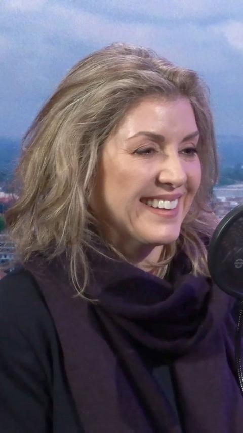 Penny Mordaunt sits in a radio studio smiling. She has long blonde hair and is sitting in front of a blue background wearing a dark purple top and scarf