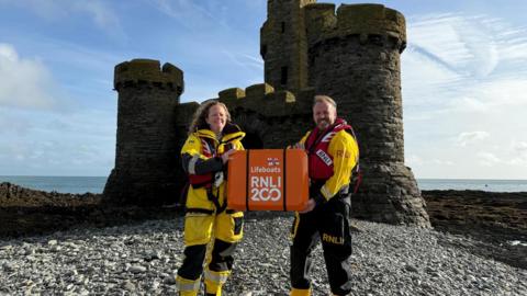 Two RNLI crew, a man and a woman, wearing their protective yellow suits and red lifejackets. They are both holding onto an orange fiberglass case with the writing "RNLI 200" on the side, which contains the scroll. They are standing in front of a small, castle-like building on an island in the sea. 