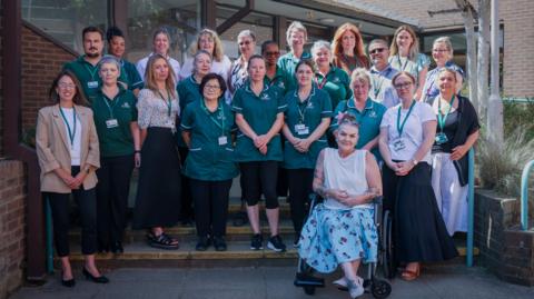 A large group of West 1 Reablement Service staff stand outside a building with one wheelchair user seated at the front of the group. Several staff members are wearing green Surrey-branded uniforms and identification badges.