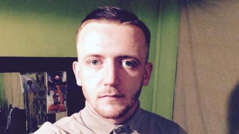 Jamie Docherty is staring at the camera. He has short, slicked back hair and is wearing a brown polo shirt with the top button fastened