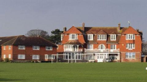 Newlands School in Seaford, a large red brick building with white windows, doors and balconies. The school also has two football goals in front of it as well as a green field.