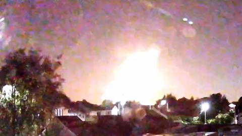 A bright flash in the night sky over some houses and gardens.