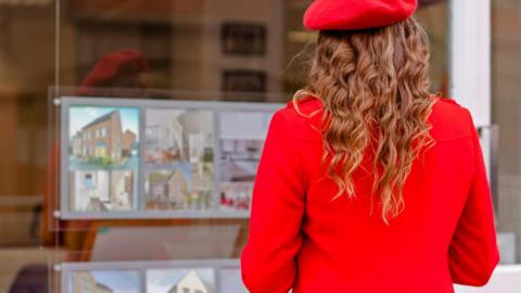 Woman wearing bright red beret and red coat faces an estate agent's window advertising homes for sale