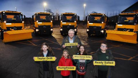 Four children stand in a car park holding registration plates reading: Taylor Drift; Humpfreeze; Who Dares Grits; and Ready Salted. With them is Councillor Barry Durkin, who is holding a plate reading: Ice-A-Saurus Rex. Behind them stand five bright yellow gritting lorries.