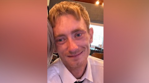 Conor Kinlan is a man with blonde hair, wearing a white, collared shirt and a black tie. He is looking directly at the camera and is smiling.