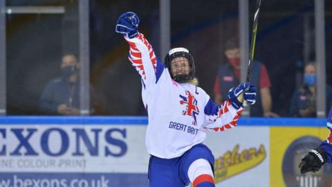 A woman in a white ice hockey jersey with Great Britain and a stylised lion printed on the front, wearing a blue helmet and holding an ice hockey stick in her left hand. She has one hand in the air and there are advertising hoardings behind her