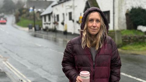 Local councillor Claire Critchison standing alongside a road, with her coat hood up while it rains