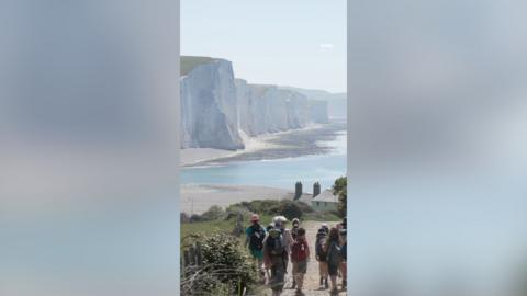 A group of people walk along a coastal path with white cliffs in the background