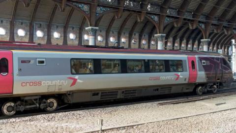 A CrossCountry train at York station, which has an arched roof. The train is silver with red accents and displays the "CrossCountry" logo. The station features decorative architectural elements and multiple windows that allow natural light in. Train tracks are visible in the foreground.