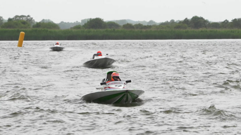 Three powerboats whizz towards the camera on a choppy river, with marsh and trees in the background. The three drivers wear orange helmets.