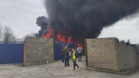 A thick black cloud of smoke above a fire burning behind a breeze block wall. The fire looks to be on top of a scrap heap of cars, with a blue burnt out car on top.
