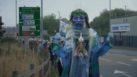 A parade of people, wearing blue and green, can be seen marching in single file along a pavement with a large road sign in the background and a building surrounded by security fencing.