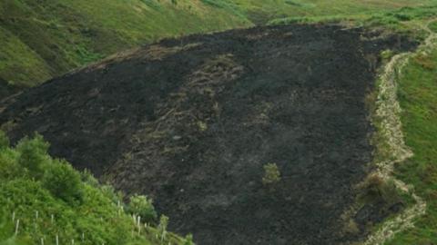 A large hillside landscape, most of which is covered in green grass and low vegetation. Cutting across the centre of the scene is a broad, dark blackened area that appears to be scorched ground.
The burned section stretches from the lower part of the valley up toward the crest of the hill. The surface within the blackened area looks uneven and textured, with patches of brown vegetation visible among the darker ground. The burned area occupies a significant portion of the hillside and has an irregular shape.
