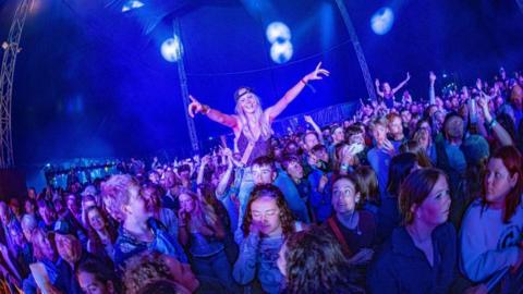 A packed outdoor audience gathers in front of a stage as the sun sets, with people raising their hands and enjoying the live music under colourful lights.