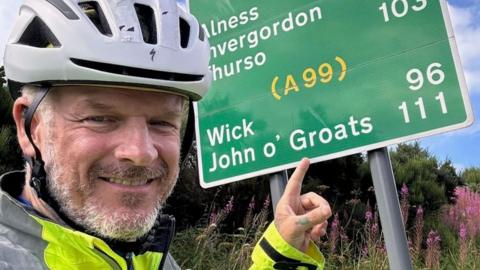 Mathew Pritchard points to a road sign that says John o'Groats. 