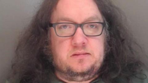 Mugshot of Madgin. He has a fat face with a scruffy grey goatee beard and thick shoulder length frizzy dark hair. He is also wearing glasses.