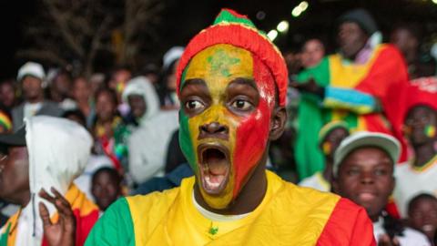 A man with his face painted in Senegal colours looks shocked with his mouth wide open.