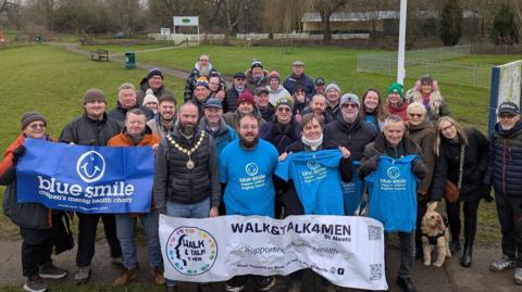 A group of people stood outside in a park. They are looking up at the camera and smiling. Those on the front row are holding blue signs or T-shirts with Blue Smile on the front, along with a blue smiley face. A white sign with Walk & Talk 4 Men St Neots is also being held at the front of the group. There are both men and women - and a dog - included in the crowd of people. 